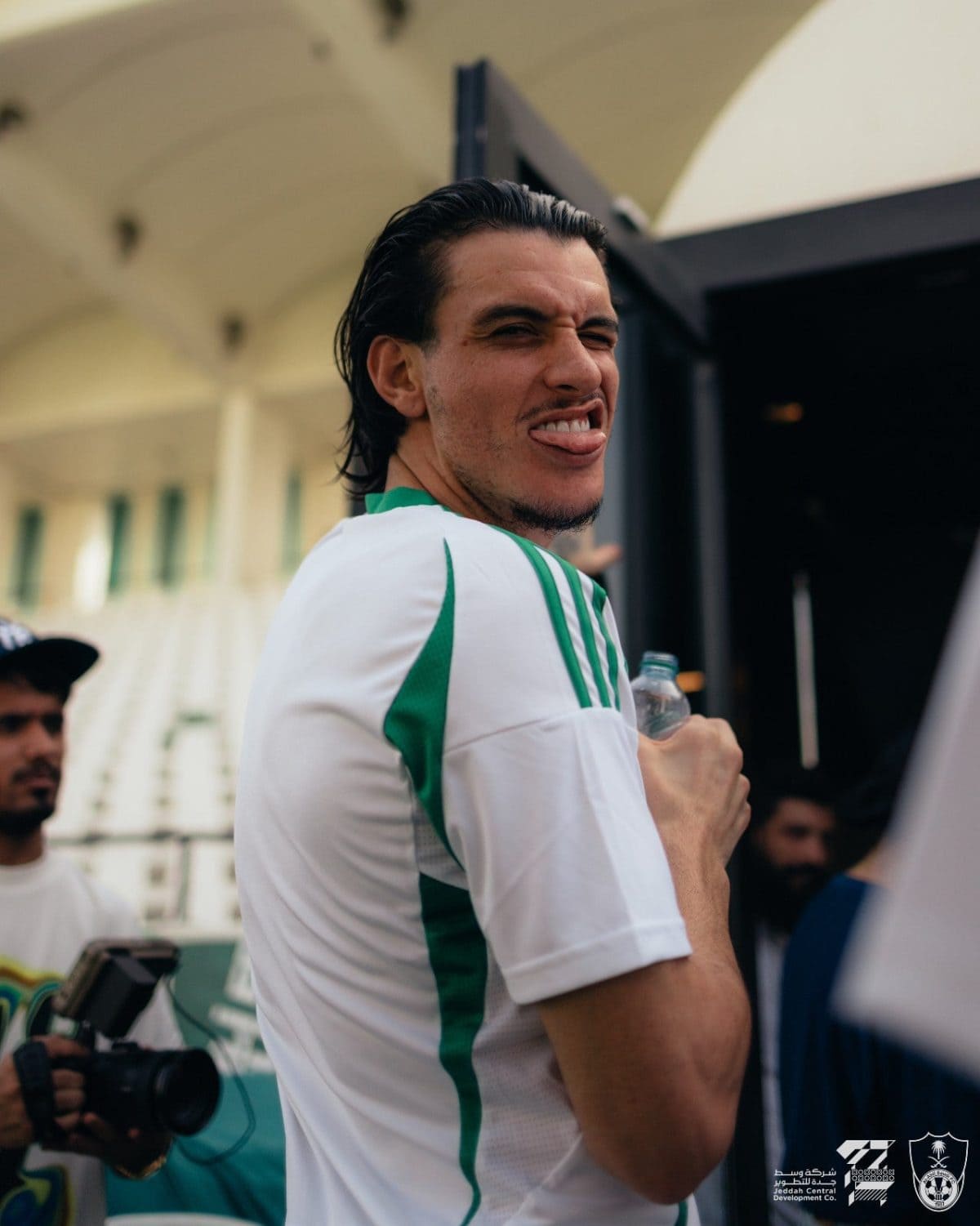 Man in a white-green sports jersey playfully sticking out his tongue while holding a water bottle at a sports event.