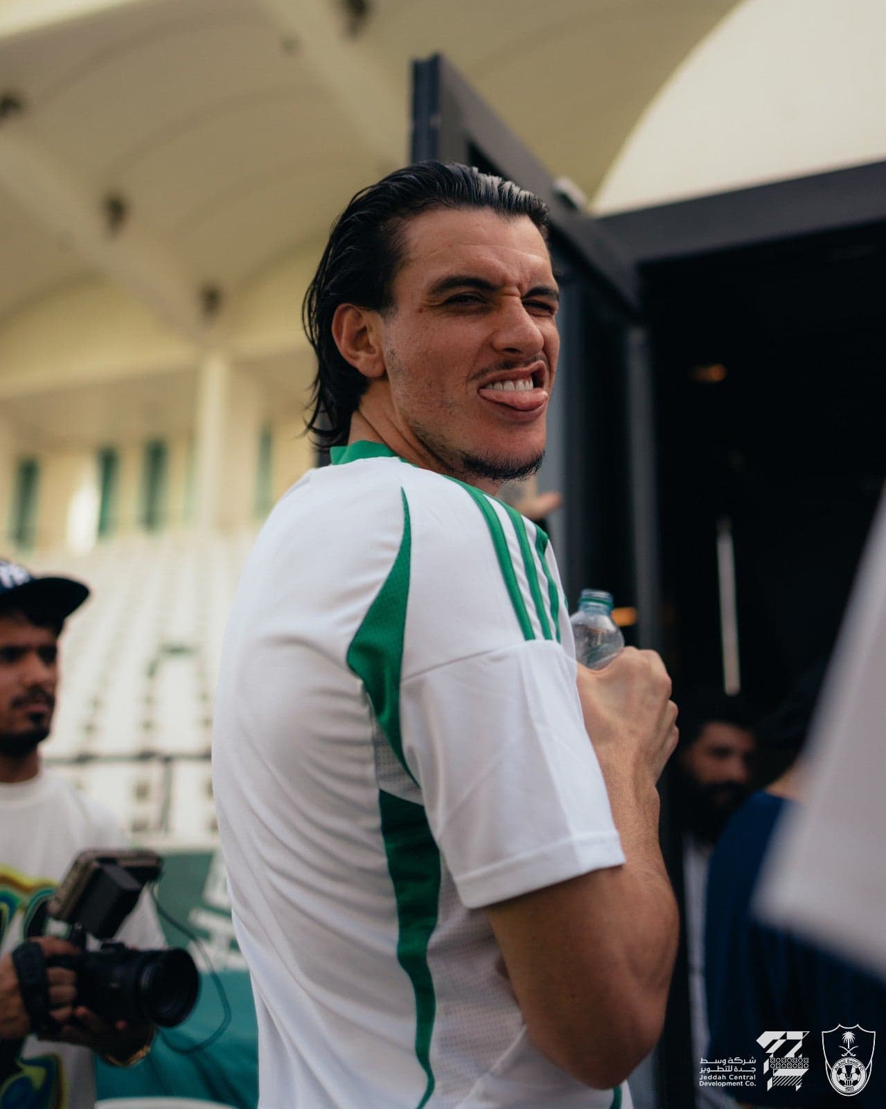 8ZGKnCAd4FgvJD3 | UAE SPORT STORE Man in a white-green sports jersey playfully sticking out his tongue while holding a water bottle at a sports event.