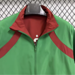 Green windbreaker with red collar and red diagonal panels, front zipper, hanging on a black hanger against a metal grid background.