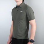 Man wearing an olive green Nike polo shirt with a white swoosh, standing against a light wall, left wrist visible with a watch.