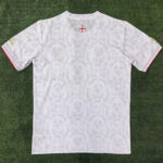 White patterned T-shirt laid flat on grass, back side up, with a small cross crest near the neckline.]','White back-view of a white short-sleeve shirt with a subtle damask pattern on green turf.