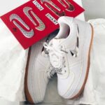 White high-top sneakers with metallic silver accents, placed on white tissue with a red display card nearby.