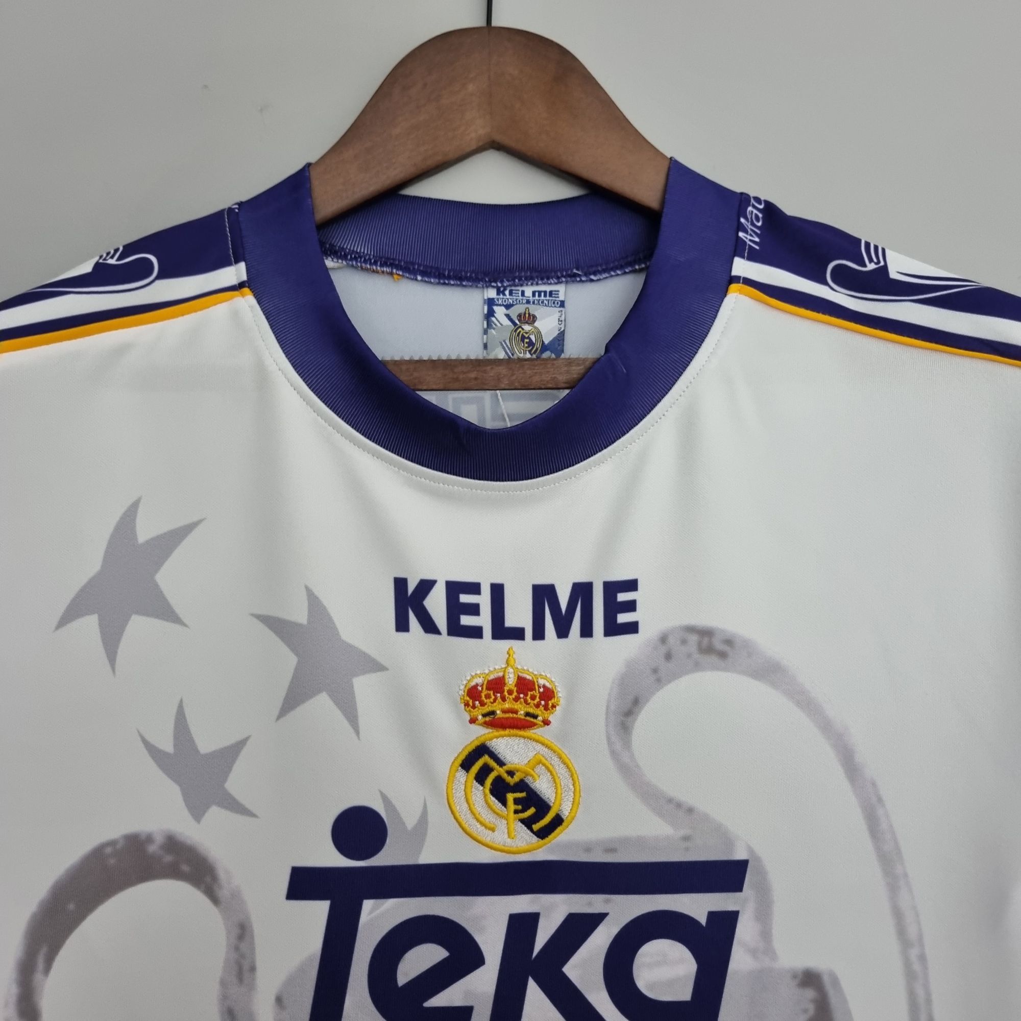 8dccd430_d05cbb00-5b9d-41da-b6b8-2412caccd5d6 | UAE SPORT STORE White Real Madrid football jersey on a wooden hanger, featuring purple trim and the club crest with a Kelme label visible in the collar area.