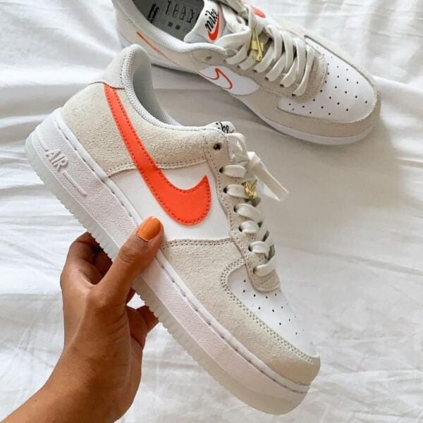 White Nike Air Force 1 sneaker with bright orange swoosh being held, another pair visible in the background on a white surface.