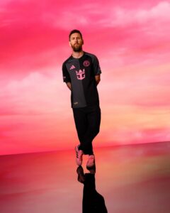 Bearded man in a black athletic jersey stands with hands behind his back on a pink gradient backdrop, reflection visible.