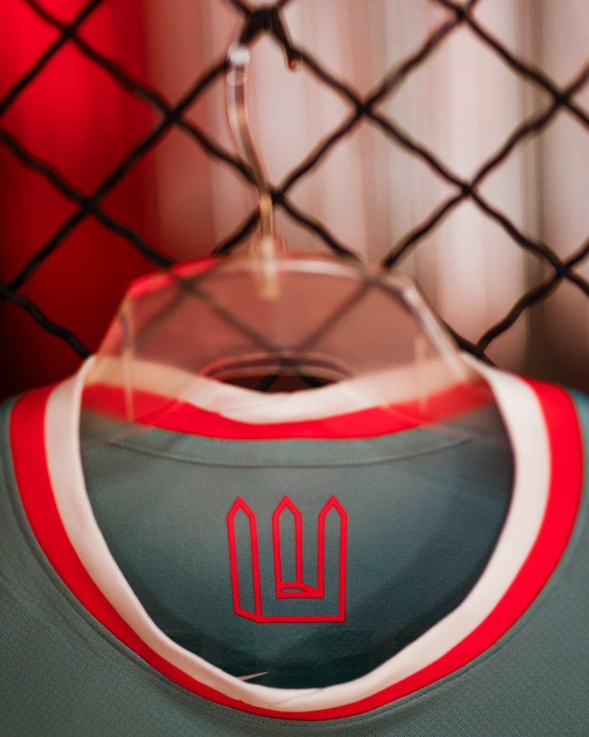 Close-up of a teal athletic jersey on a plastic hanger, with red and white trim, hanging in front of a chain-link fence; a red geometric logo is on the chest area.