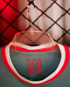 Close-up of a teal athletic jersey on a plastic hanger, with red and white trim, hanging in front of a chain-link fence; a red geometric logo is on the chest area.