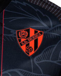 Shield-shaped red and black crest of S.D. Huesca with a volleyball emblem, on dark patterned fabric (sports jersey).