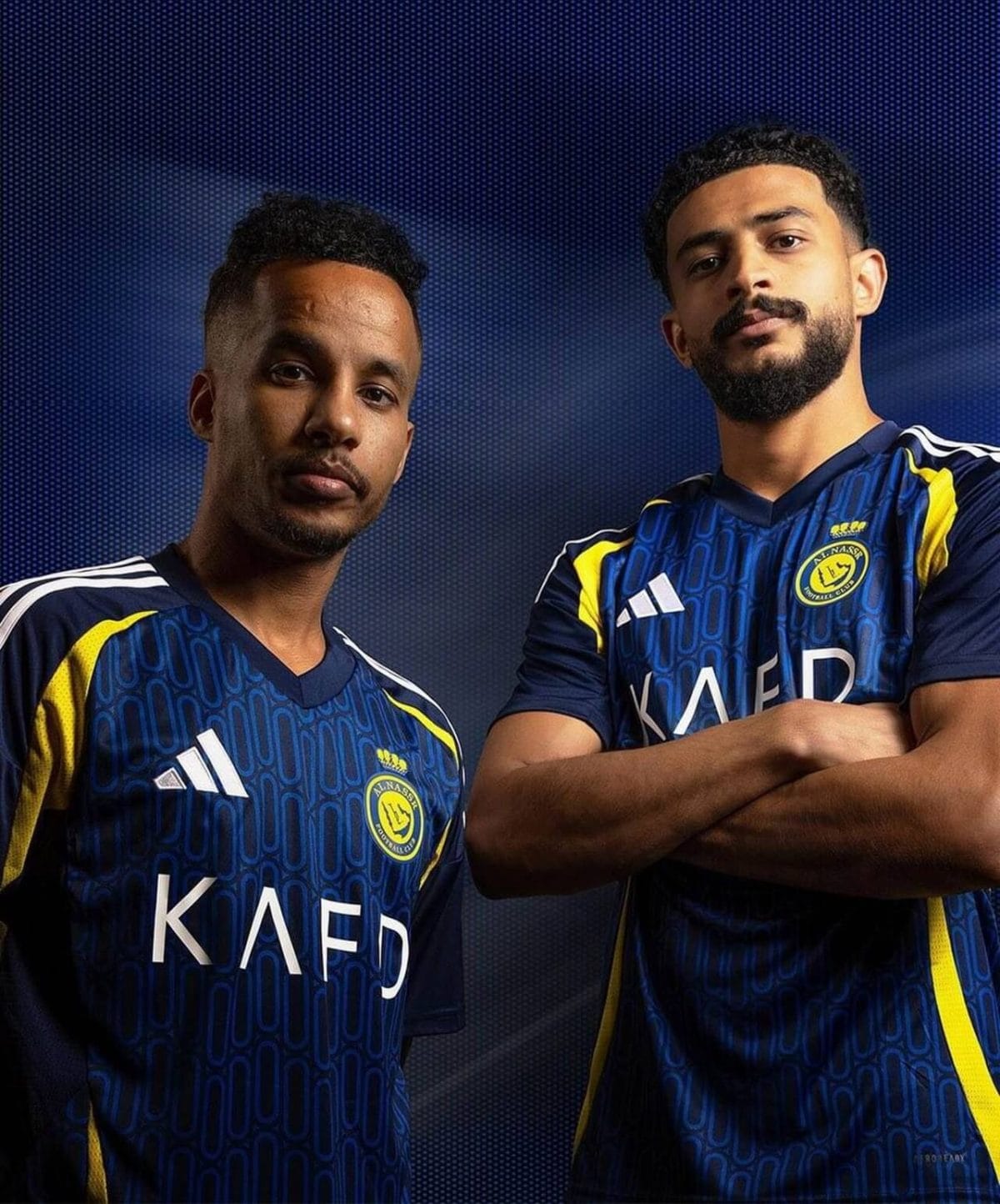Two young men in matching blue athletic jerseys pose confidently against a dark, dotted blue background, arms folded.