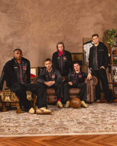 Five men in matching black-and-red striped jackets pose in a vintage living room, sitting and standing around a leather sofa with retro decor.