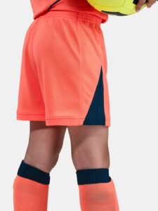 Side view of bright coral athletic shorts with a navy side panel and vent seam, paired with navy knee-high socks. A yellow ball is held nearby.