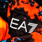 Close-up of black fabric with vivid orange and blue abstract pattern and a white 'EA' logo with horizontal stripes.