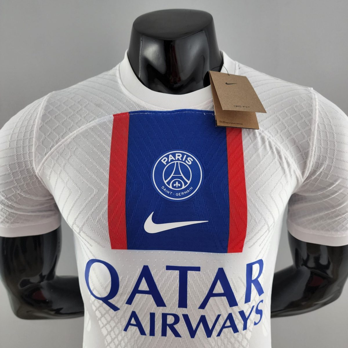 Mannequin wearing a white Paris Saint‑Germain jersey with blue and red stripes and a brown tag attached to the collar area, front view.