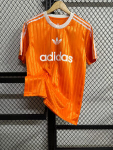 Bright orange Adidas T-shirt with white stripes on the shoulders and trefoil logo on a wooden hanger against a mesh background.