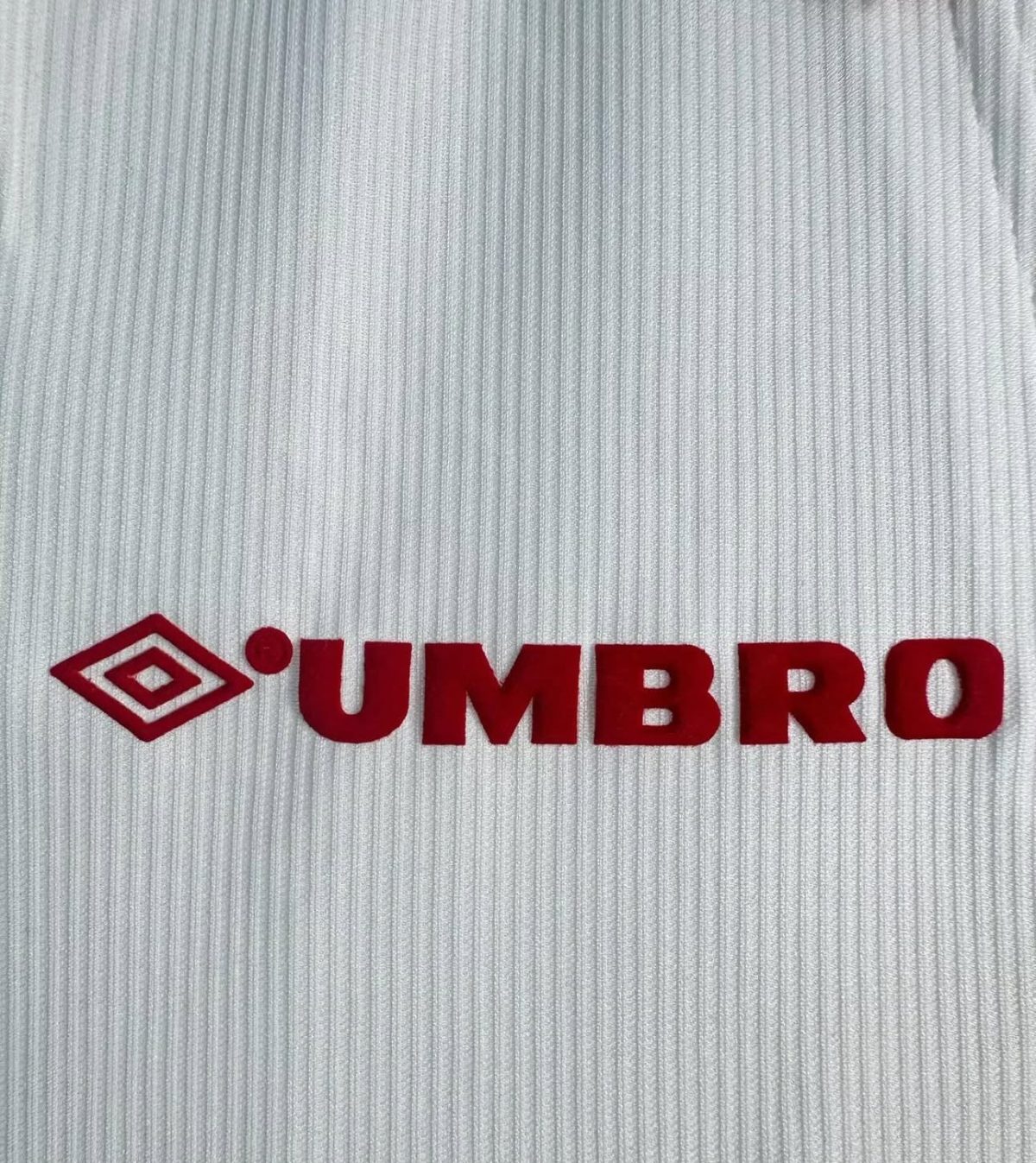 Red Umbro wordmark with the diamond emblem printed on white ribbed fabric (close-up). Readable branding on sports material.