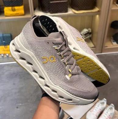 Gray running shoe with a gold logo, held up in a store with shelves in the background, showing a chunky white midsole and yellow outsole edge.