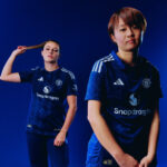 Two female athletes pose in blue Manchester United soccer jerseys against a dark blue background, Snapdragon logo visible on fronts.