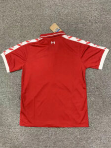 Back view of a red polo shirt laid flat on a gray carpet, white stripes along the shoulders and white sleeve cuffs, tag at the collar.