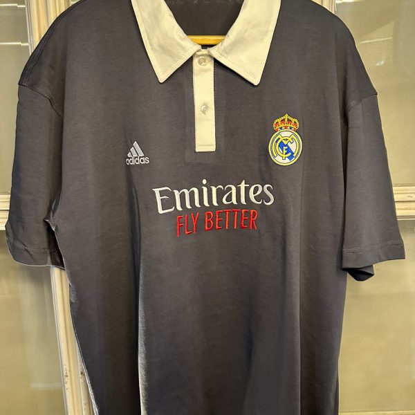 Dark gray Real Madrid polo shirt on a hanger, white collar, Adidas logo on the left, Real Madrid crest on the right, Emirates Fly Better sponsor across the chest.