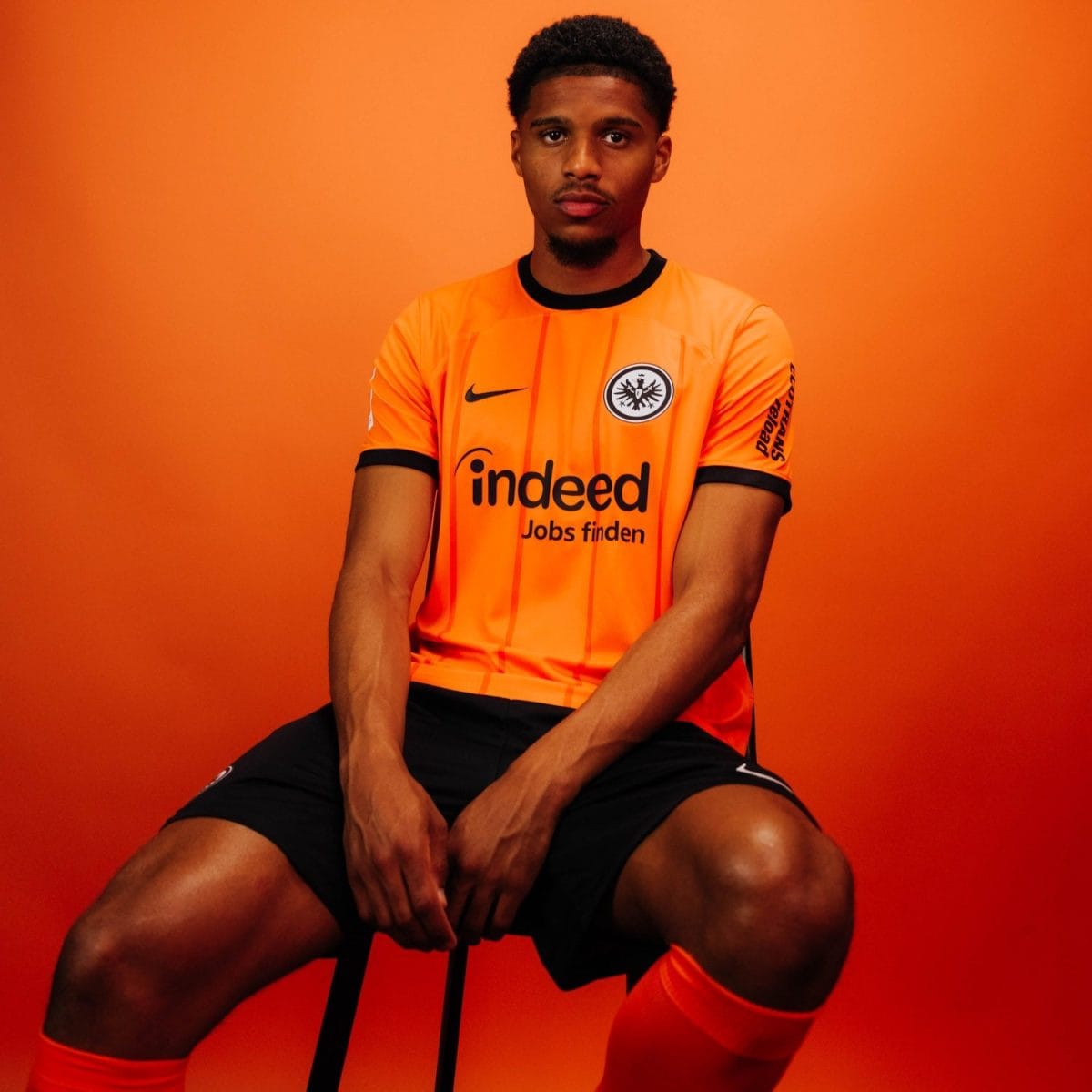 Portrait of a young man in a bright orange soccer jersey seated against an orange background, arms resting on his knees, facing the camera. The jersey features a crest on the chest and the word 'indeed' as a sponsor.