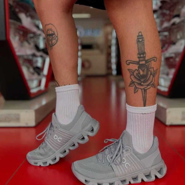Person standing in a store wearing gray athletic sneakers and white socks; left calf has a circular tattoo, right calf has a dagger and rose tattoo.
