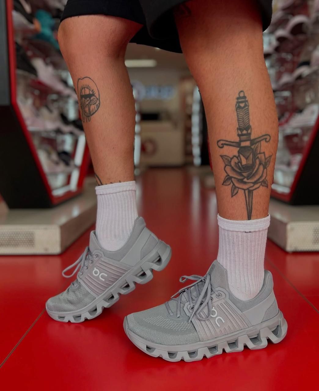 93b251e2-e8e3-4c60-b22b-fe209c4253a9 | UAE SPORT STORE Person standing in a store wearing gray athletic sneakers and white socks; left calf has a circular tattoo, right calf has a dagger and rose tattoo.