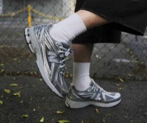 Close-up of a gray New Balance running shoe on a person’s leg with a white sock, foot raised beside a chain-link fence.