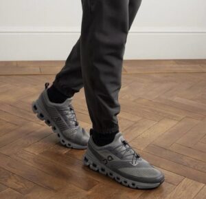 Person wearing gray athletic sneakers stepping on a wooden floor wearing black joggers.