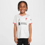Smiling young child wearing a white Liverpool FC jersey with red trim and sponsor logo, standing against a light background.