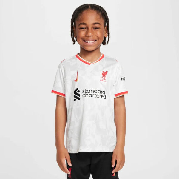 Smiling young child wearing a white Liverpool FC jersey with red trim and sponsor logo, standing against a light background.