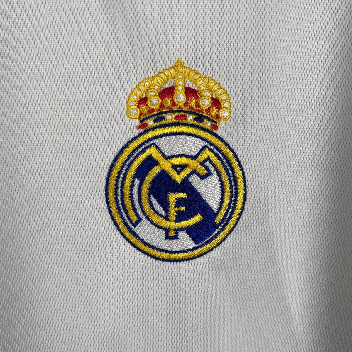 Partial Real Madrid crest embroidered in yellow, blue, and white on a white textured fabric background.