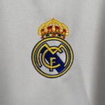 Partial Real Madrid crest embroidered in yellow, blue, and white on a white textured fabric background.