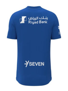 Back view of a blue T-shirt with white Riyad Bank logo on the upper back and a 'SEVEN' logo near the lower back.