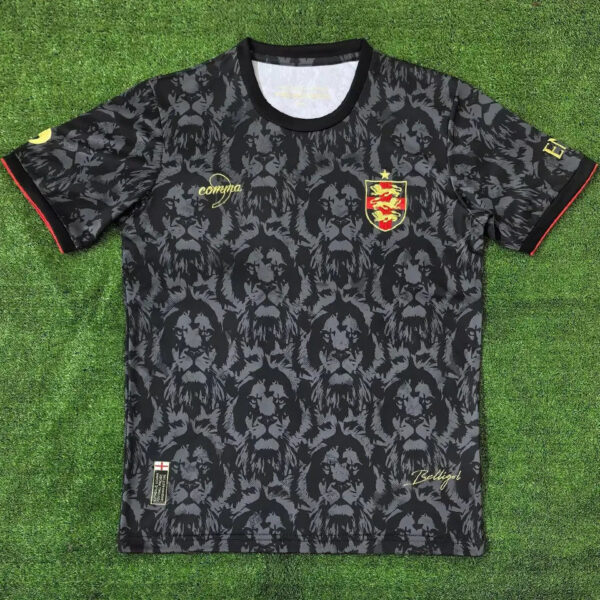 Dark gray/black lion-patterned soccer jersey with a gold crest on the left chest and gold script branding on the right, red-trimmed sleeves, and a small England flag tag near the hem bottom left.