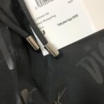 Close-up of black fabric with Dior-branded metal zipper pulls and a white tag showing barcode and 'Toile jean logo DIOR' text, indicating a Dior garment