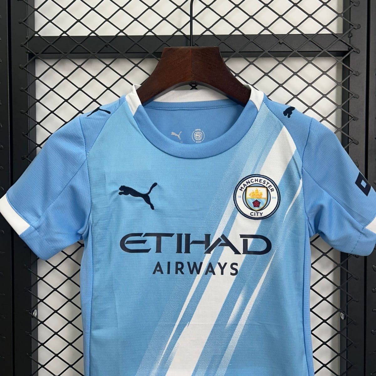 Blue Manchester City football jersey on a wooden hanger in front of a metal grid locker, logo visible on chest area.