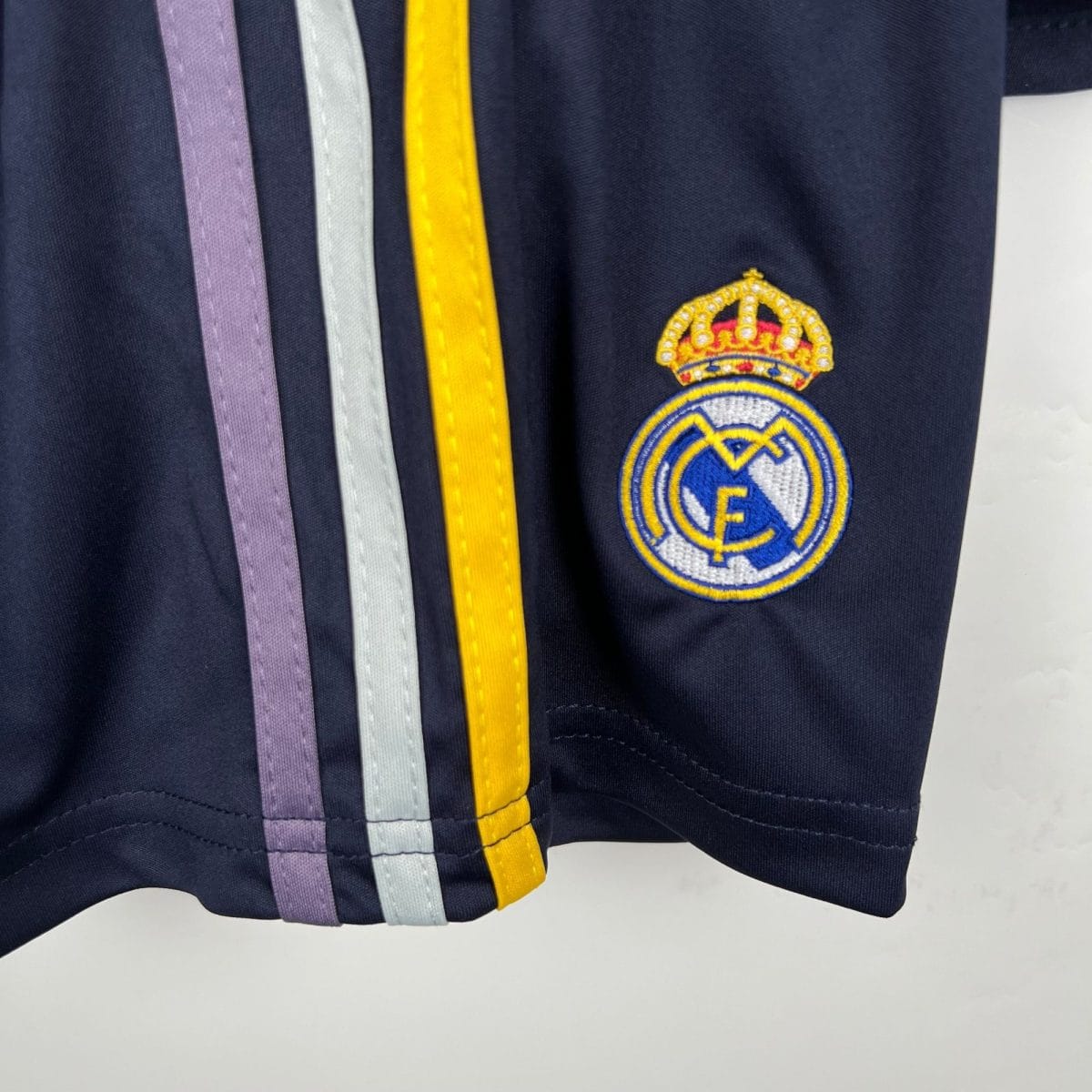 Close-up of dark fabric with five narrow vertical ribbons (purple, light purple, light gray, yellow) and a circular embroidered crest featuring a crown on the right edge.
