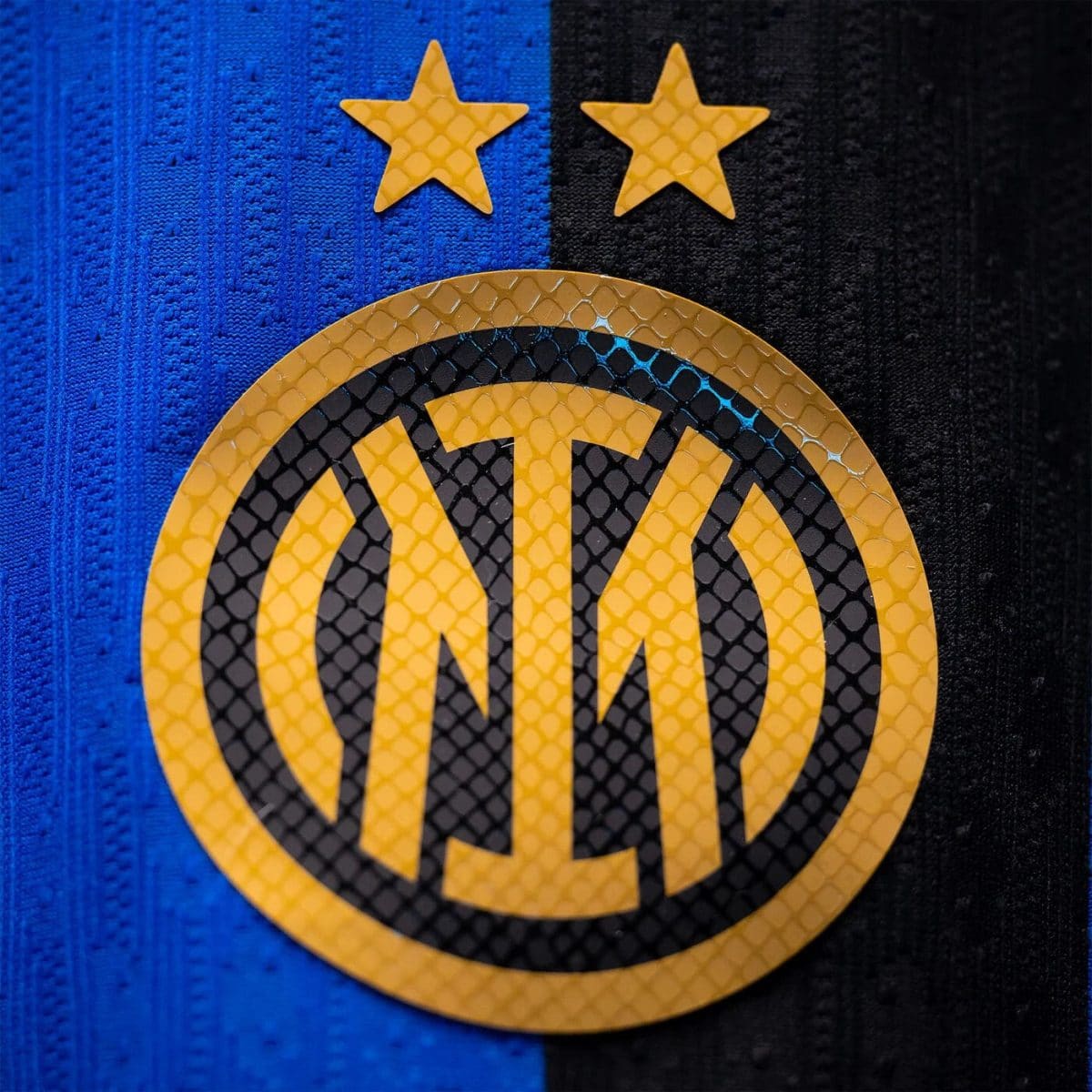 Close-up of a yellow circular emblem with a black M on a blue and black striped jersey, with two gold stars above.
