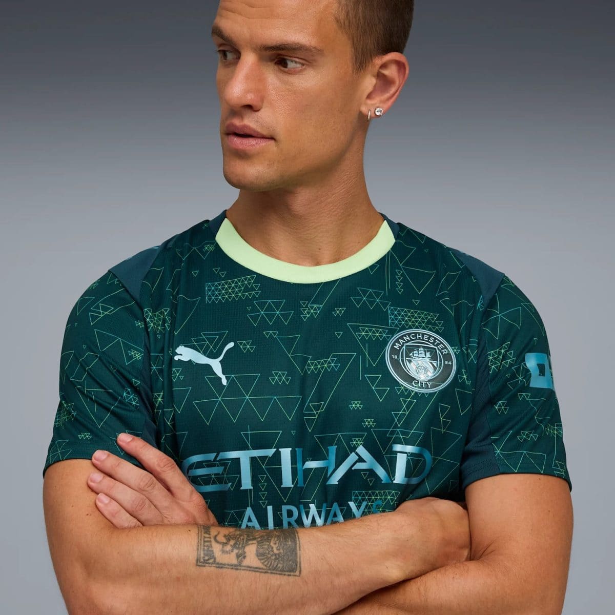 Man wearing a Manchester City football jersey with geometric teal pattern, mint collar, crest visible, arms crossed, looking to the left.