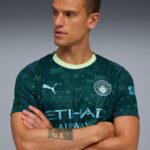 Man wearing a Manchester City football jersey with geometric teal pattern, mint collar, crest visible, arms crossed, looking to the left.