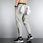 Back view of a person wearing light gray tapered joggers with black cuffs, a white shirt tied at the waist, and black Nike sneakers with a white sole; a black wristwatch is on the left arm.
