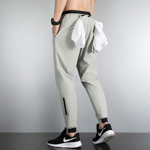 Back view of a person wearing light gray tapered joggers with black cuffs, a white shirt tied at the waist, and black Nike sneakers with a white sole; a black wristwatch is on the left arm.