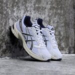 Pair of white and navy ASICS running shoes with mesh panels leaning against a textured concrete wall.