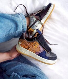 Colorful patchwork sneaker with brown laces held over blue jeans on a white surface.