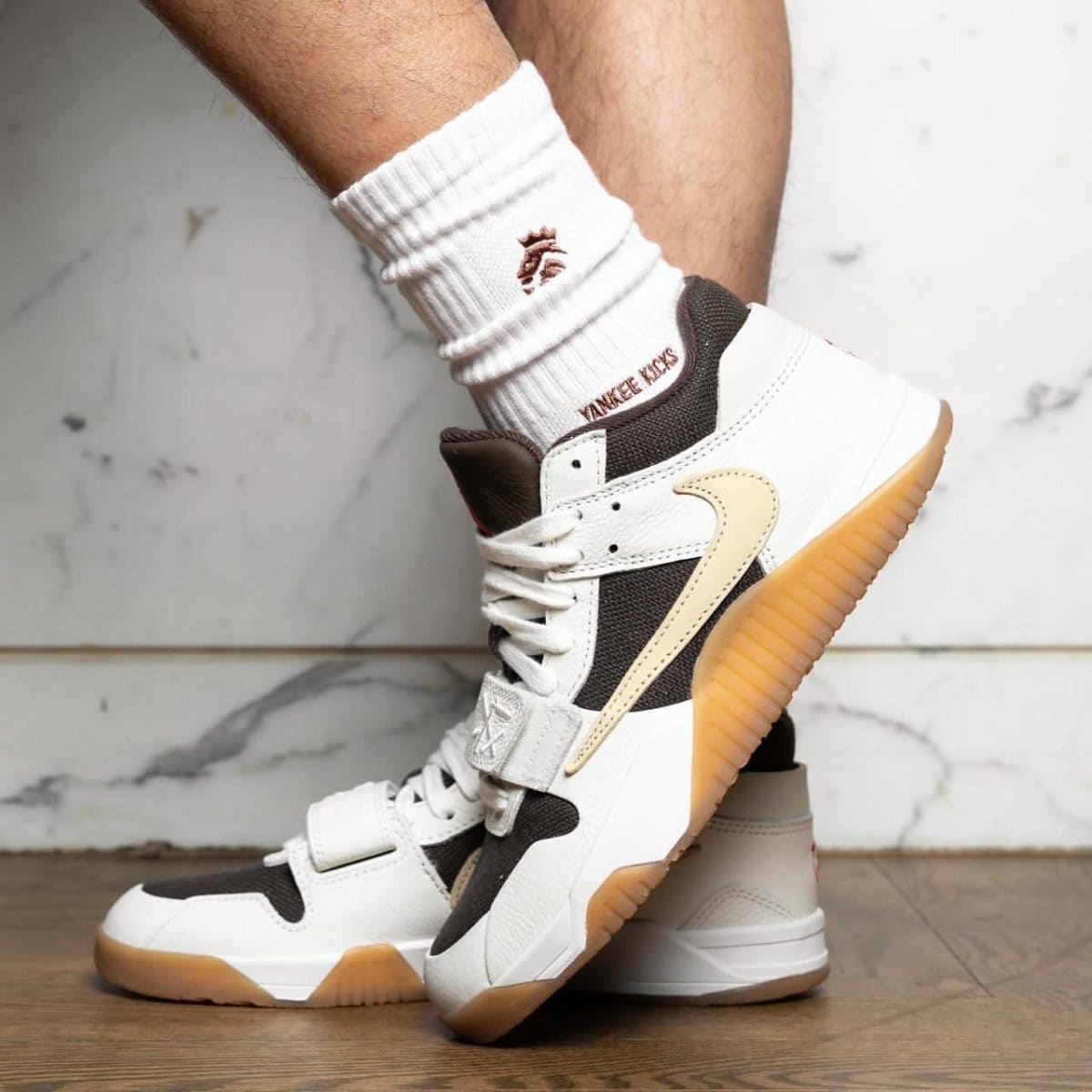 White and black sneakers with a tan gum sole, shown mid-step on a wooden floor with a white sock partially covering the ankle.