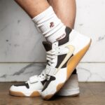 White and black sneakers with a tan gum sole, shown mid-step on a wooden floor with a white sock partially covering the ankle.
