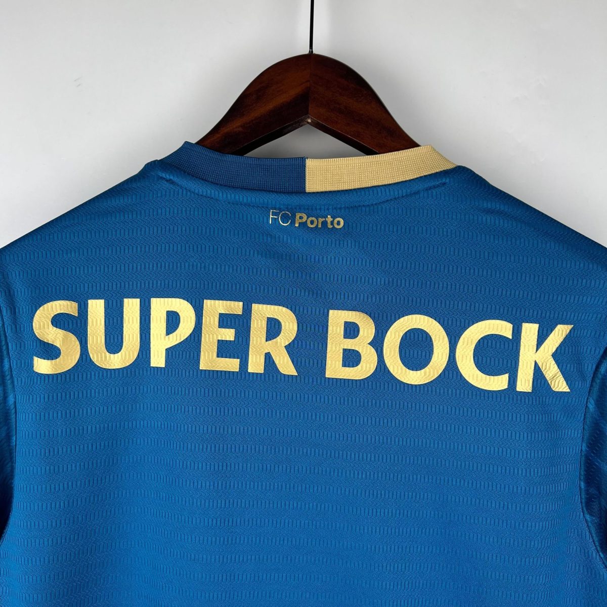 Blue FC Porto sports jersey hanging on a wooden hanger; gold lettering reads 'SUPER BOCK' across the chest and a light beige collar detail is visible.