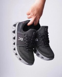9940bb22-bc76-4c75-a569-9bcdca8d23ae | UAE SPORT STORE Hand holding a pair of black athletic running shoes with thick grey soles, brand logos visible on the sides and tongue.