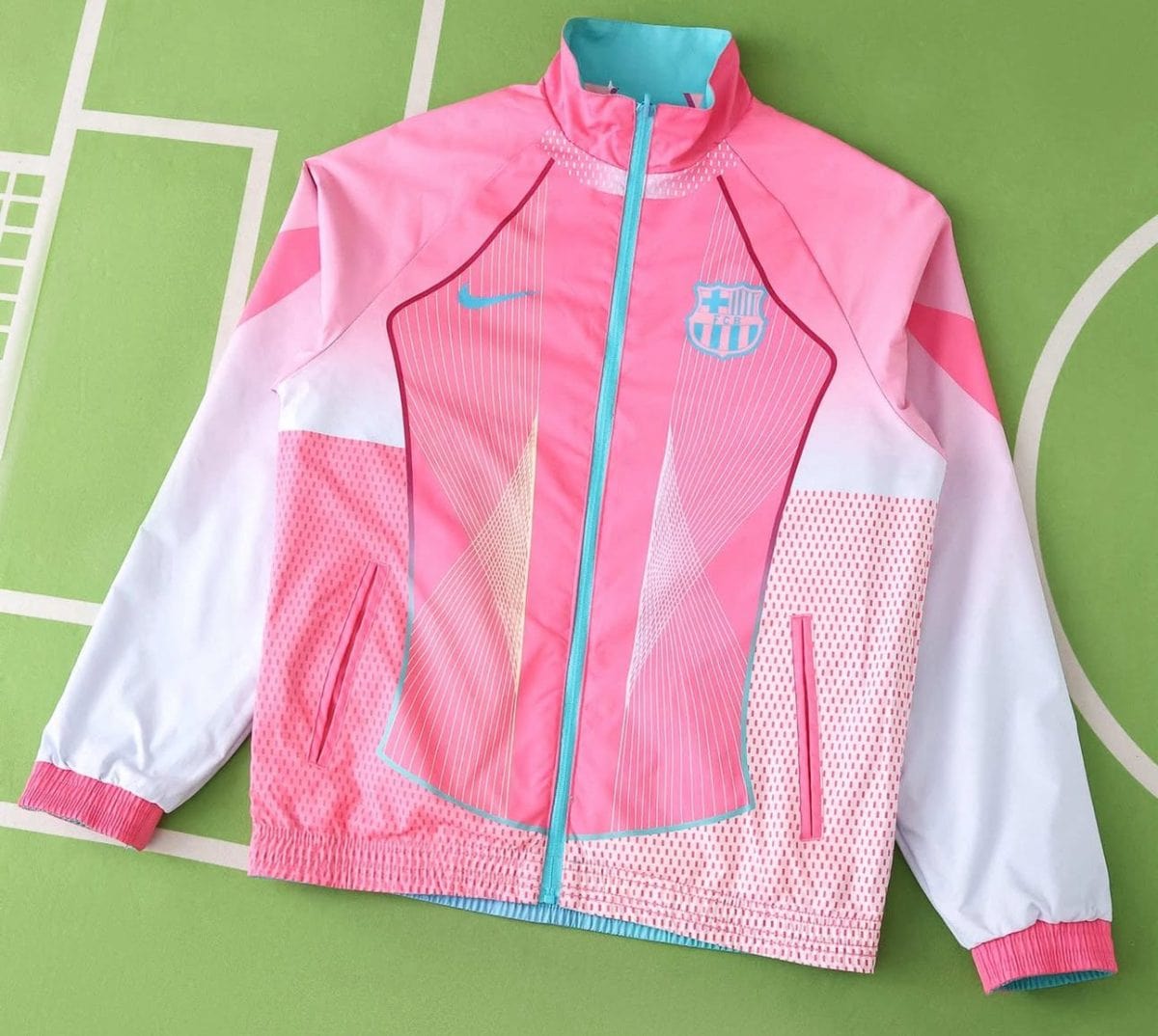 Pink Barcelona windbreaker with teal zipper and crest on the left chest, white sleeves, and geometric teal accents against a green background.