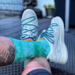 Person sitting outdoors with legs crossed, teal tie-dye socks and white sneakers with teal laces, tattoo on leg visible on a woven table surface nearby